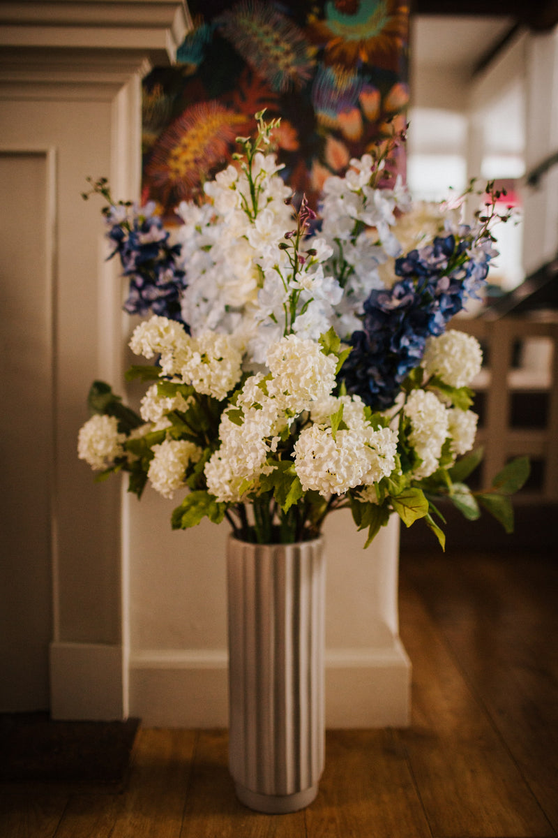 Luxury artificial delphinium, viburnum Snowball and salal foliage expertly arranged by our team of florists into a ceramic vases or available as a beautifully wrapped bouquet. Perfect for corporate and larger spaces.