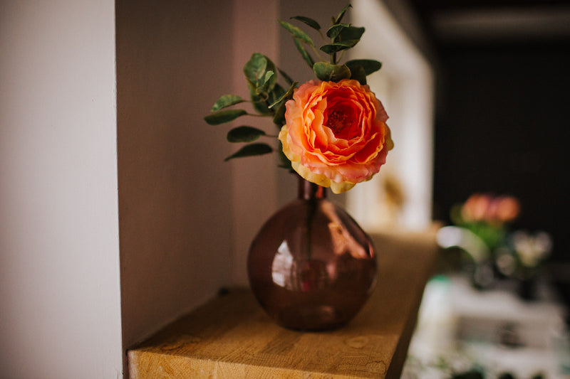 Deceptively realistic and timeless artificial ranunculus with seasonal foliage styled in a pink glass bottle vase. Perfect for bedside tables or areas in need of a touch of colour.