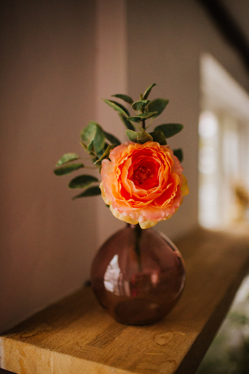 Deceptively realistic and timeless artificial ranunculus with seasonal foliage styled in a pink glass bottle vase. Perfect for bedside tables or areas in need of a touch of colour.