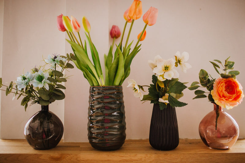 Deceptively realistic and timeless artificial ranunculus with seasonal foliage styled in a pink glass bottle vase. Perfect for bedside tables or areas in need of a touch of colour.
