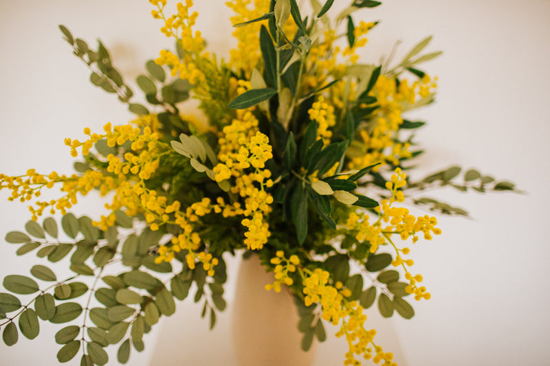 An abundance of artificial mimosa, olive branches and soft grey foliage arranged in a chic off white vase or available as a gift wrapped bouquet.  Perfect for the home or to gift.