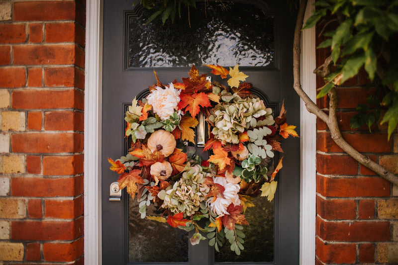 An eye catching abundance of autumnal foliage paired with silky cream and blush pink hydrangea, blooms and pumpkins in a celebration of the amazing colours and textures of the season.    This luxurious faux wreath has been created by our team of skilled florists ready to add instant warmth and comfort to any space.