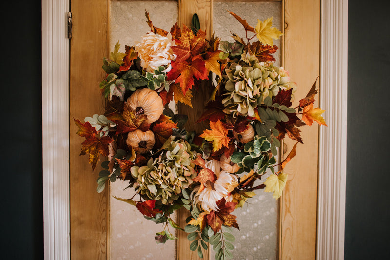An eye catching abundance of autumnal foliage paired with silky cream and blush pink hydrangea, blooms and pumpkins in a celebration of the amazing colours and textures of the season.    This luxurious faux wreath has been created by our team of skilled florists ready to add instant warmth and comfort to any space.